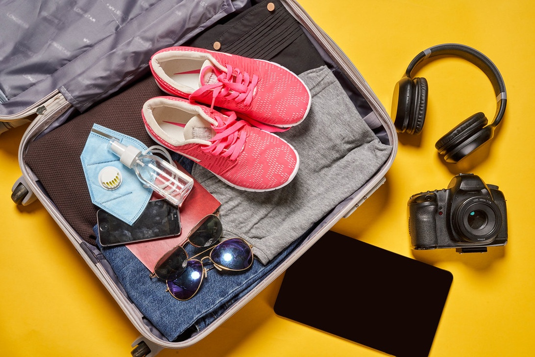 En öppen resväska på en ljusgul bakgrund innehållande prydligt arrangerade researtiklar inklusive rosa sneakers, vikta kläder, solglasögon, en ansiktsmask, handsprit och en smartphone. Bredvid resväskan finns en DSLR-kamera, hörlurar och en surfplatta.