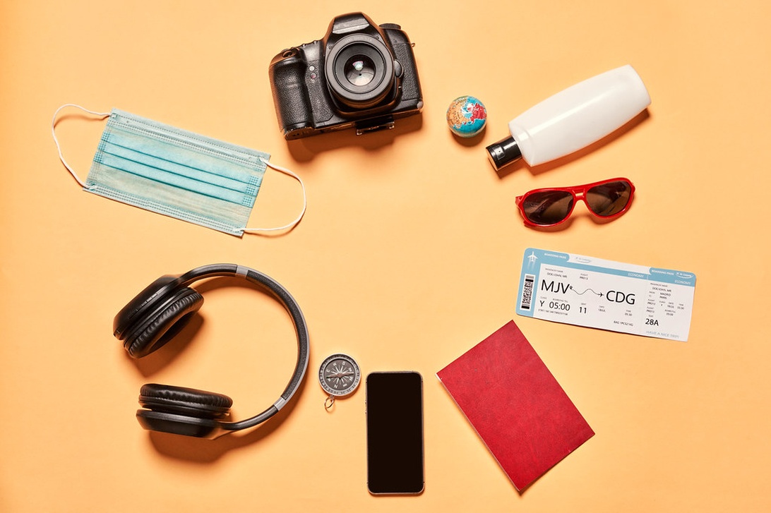 En noggrant arrangerad flatlay av reseförnödenheter på en varm orange/persikofärgad bakgrund inklusive en DSLR-kamera, hörlurar, ansiktsmask, boardingkort/flygbiljett, rött pass, liten jordglobmodell, kompass, smartphone, solglasögon och handspritflaska.