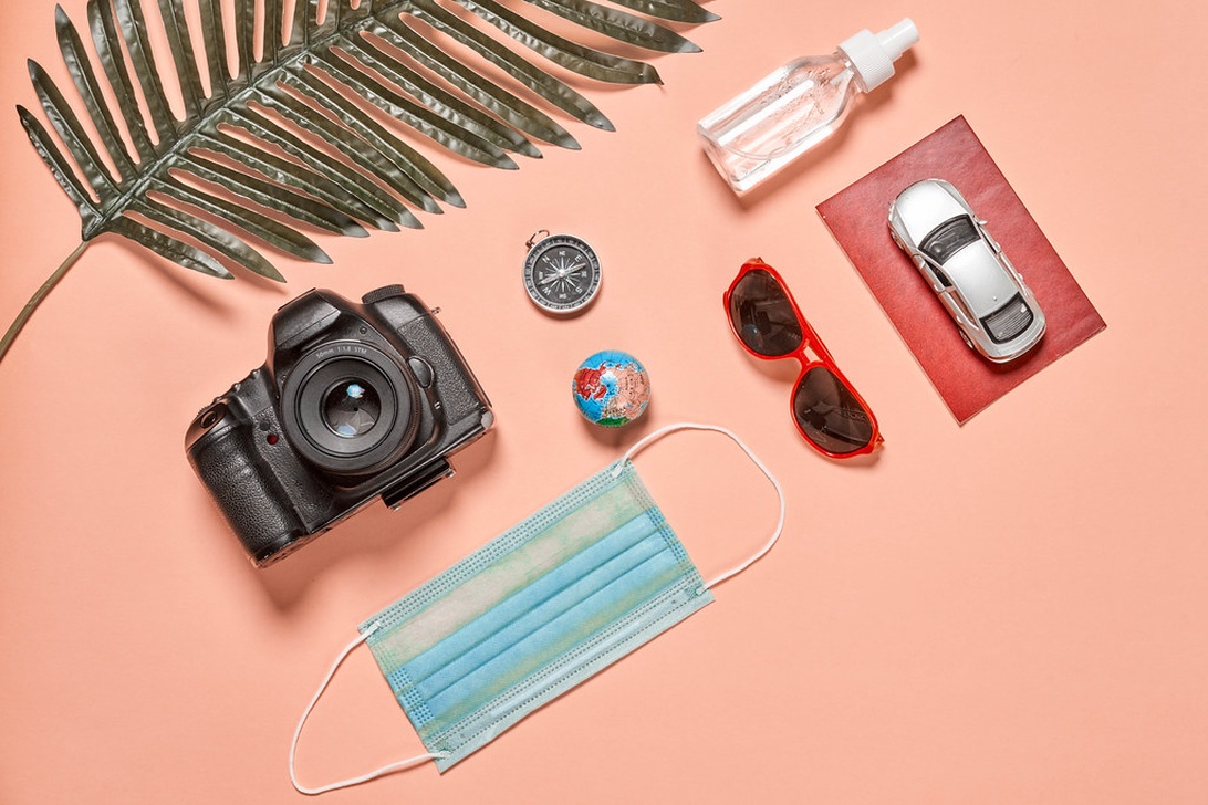 En noggrant arrangerad flatlay av reseförnödenheter på en varm orange/persikofärgad bakgrund inklusive en DSLR-kamera, hörlurar, ansiktsmask, boardingkort/flygbiljett, rött pass, liten jordglobmodell, kompass, smartphone, solglasögon och handspritflaska.